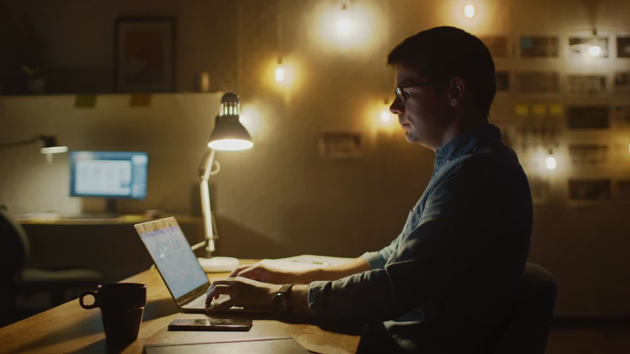 Concentrated Man Sitting at His Desk in Office Studio Working on a Laptop in the Evening. Work with Data, Analyzing Statistics. 360 Degree Tracking Arc Shot Movement. Slow Motion