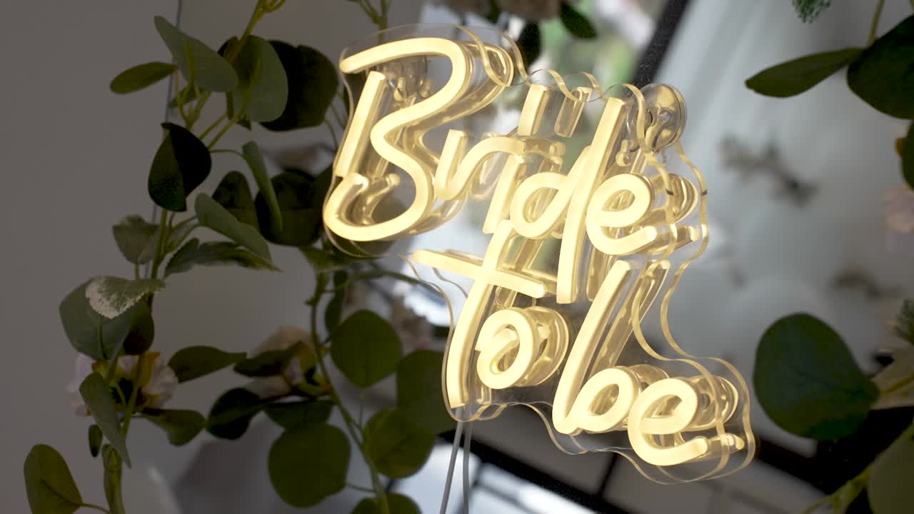 Un cartello luminoso indica una futura sposa. È posizionato tra le piante in una stanza. Questa scenografia evidenzia una celebrazione per l'imminente matrimonio. Amici e familiari si riuniscono per onorare la sposa.