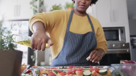 happy african american woman in apron pouring oil on vegetables in kitchen, slow motion