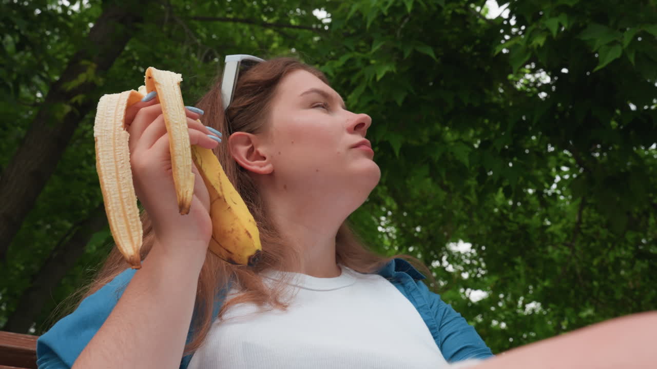푸르른 나무와 자연광으로 둘러싸여 평화로운 여름 분위기를 담아낸, 사려 깊은 표정으로 주변을 둘러보며 야외에서 플랜테인을 먹고 있는 여성의 올려다본 앵글 뷰