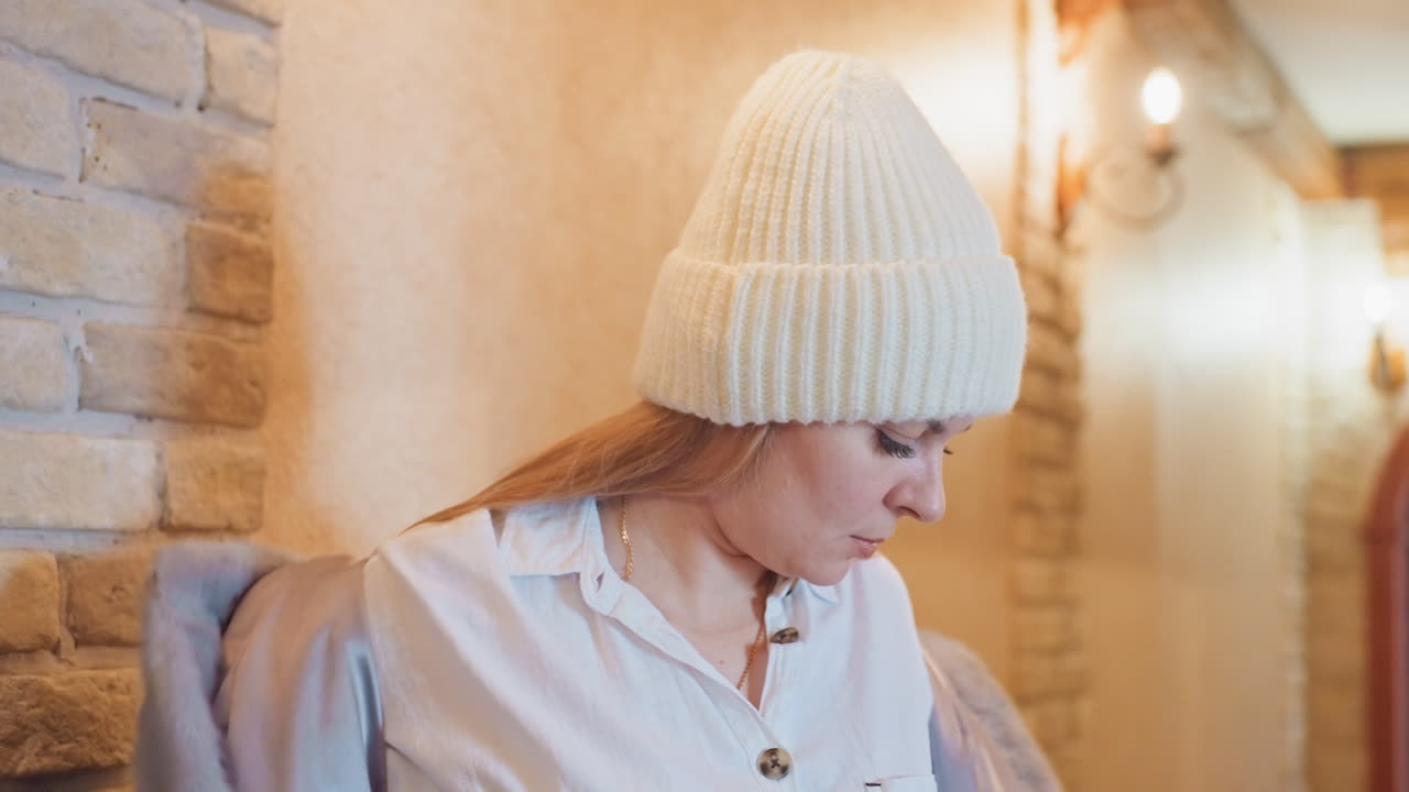 Gelassenes Porträt einer Frau in gemütlicher Umgebung. Ruhiges Bild einer Frau mit Anhänger nahe einer Backsteinwand. Friedliche Szene, die eine Frau mit Strickmütze zeigt, die ihren Mantel neben einer Backsteinwand mit nachdenklichem Blick richtet