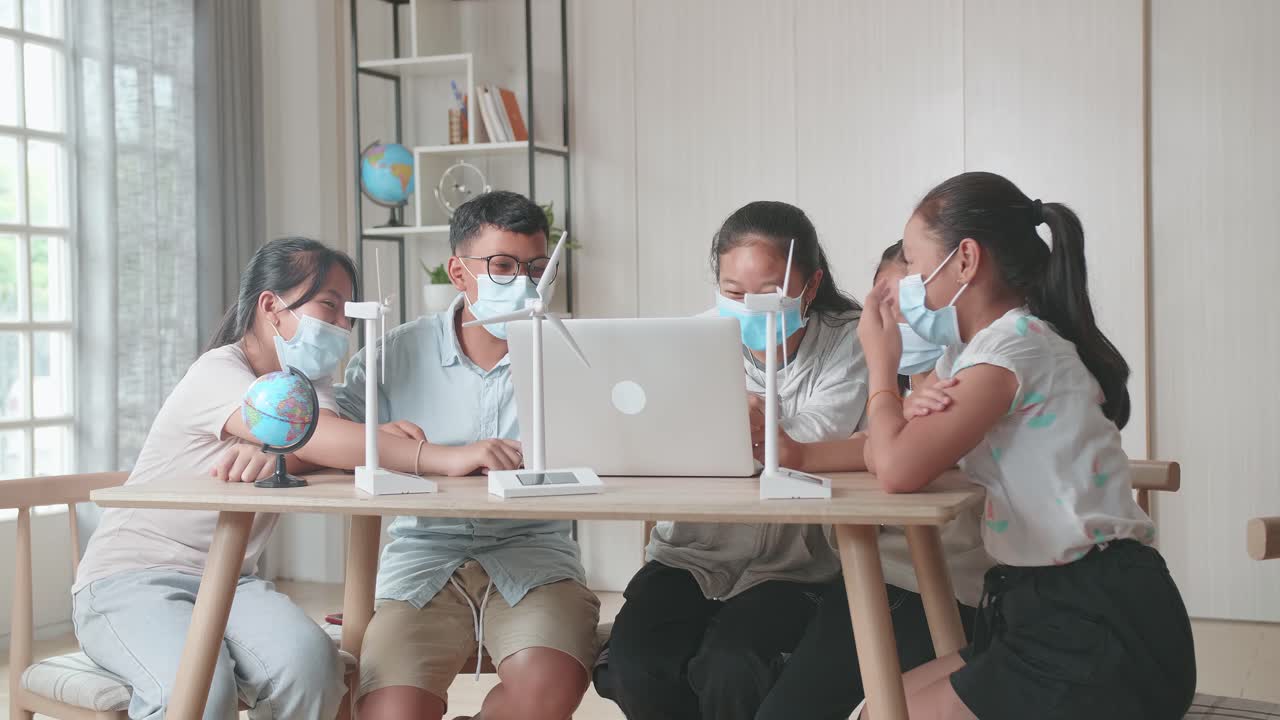 Young Asian Children Are Wearing Facemasks And Work As A Team Using Laptop Computers To Program Wind Turbines. Classroom With Kids Learning About Eco-Friendly Forms Of Renewable Energy