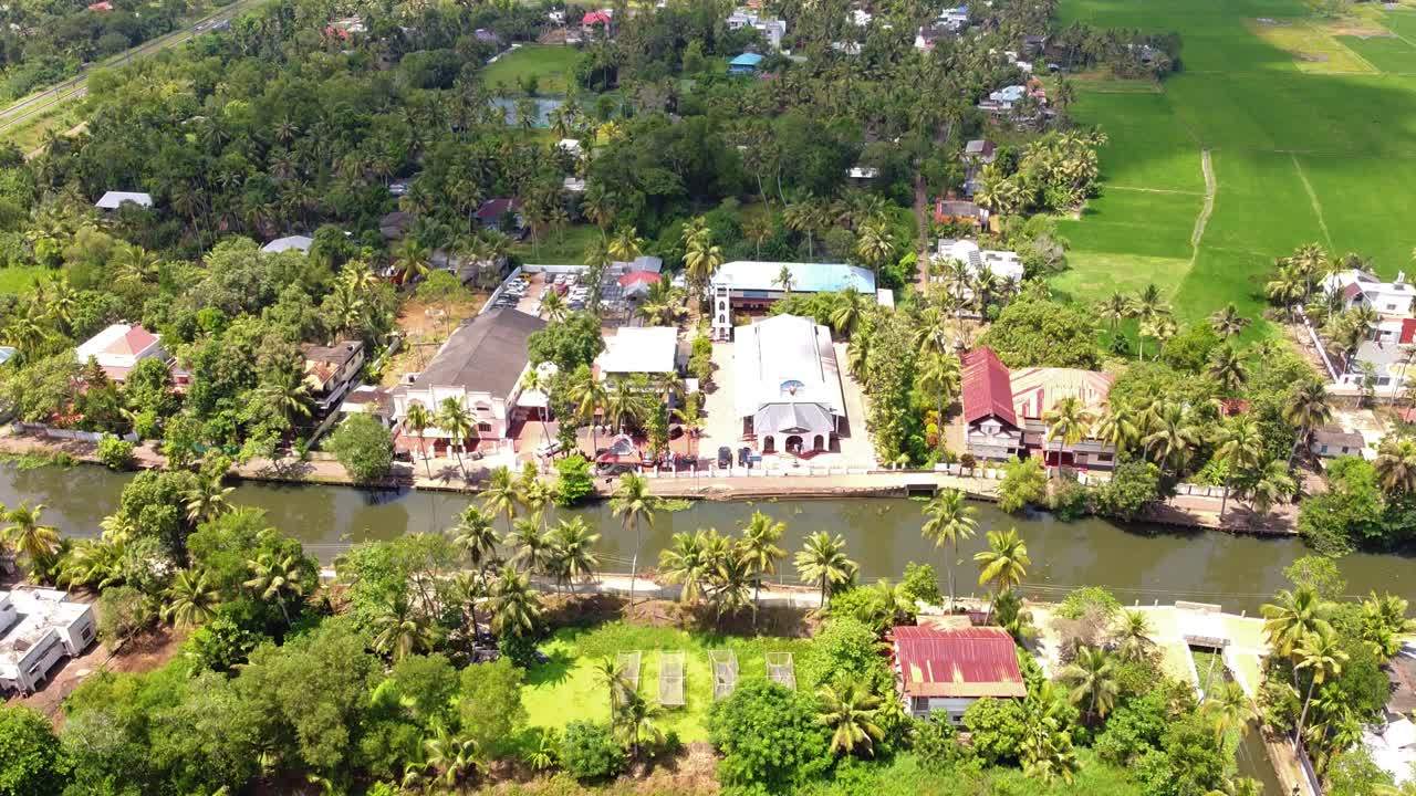 Expansive aerial view of a cluster of churches and buildings situated along a serene backwater canal, surrounded by lush tropical greenery and paddy fields in Kerala