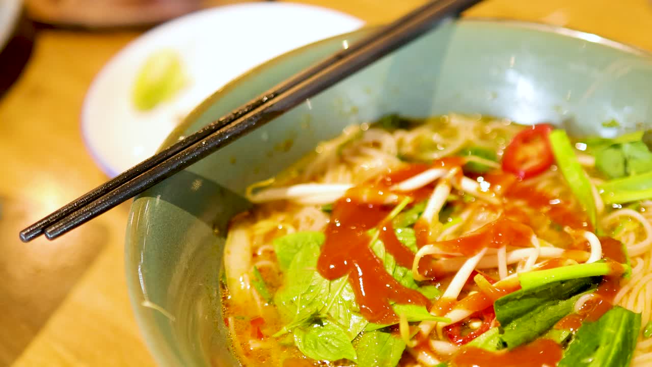 A vibrant bowl of Vietnamese pho is garnished with fresh herbs and sauce in a Bangkok restaurant, captured in warm lighting