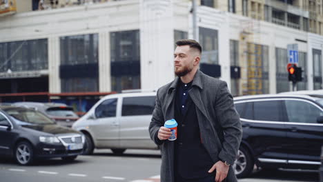 Handsome man in the city. Young businessman waiting for someone on the urban traffic background. Man in coat holds a plastic glass with coffee and looks on watch.