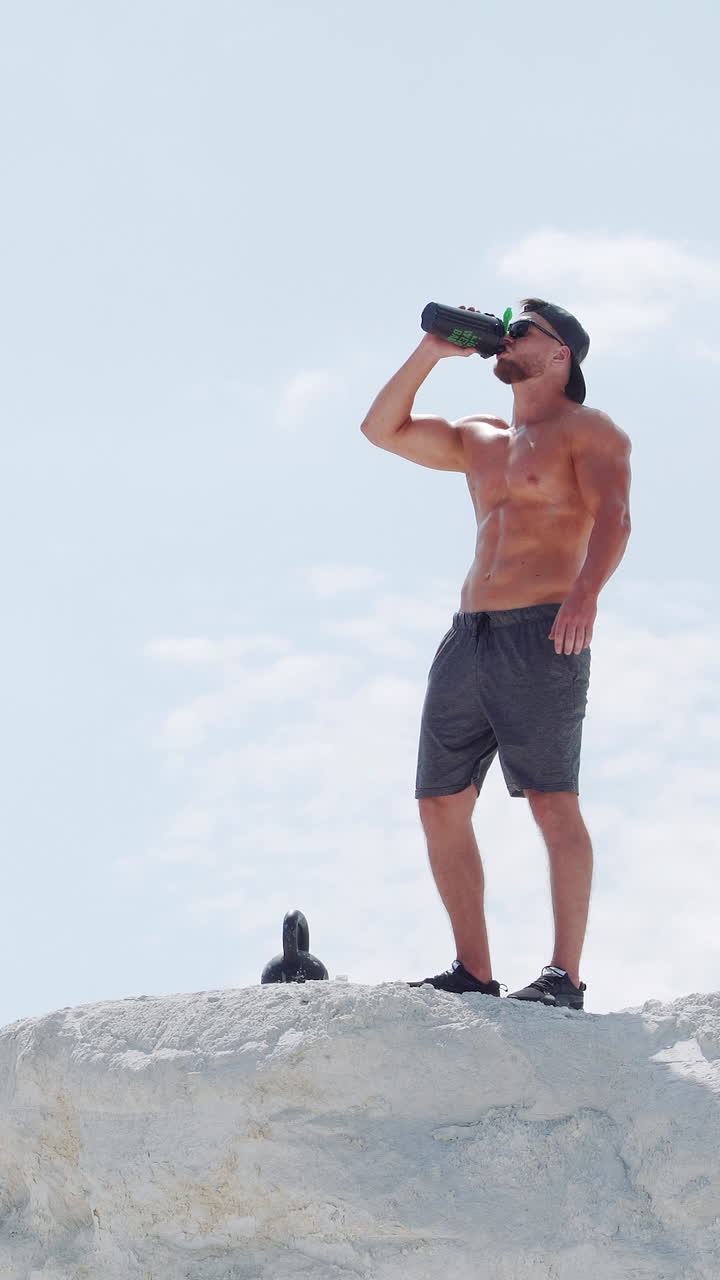 Strong man drinks water from a bottle in an outdoor training session. Athlete on a white mountain against the blue sky. Vertical video