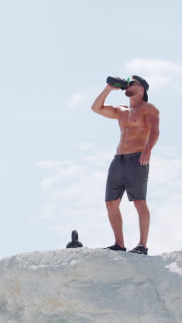 strong man drinks water from a bottle in an outdoor training session. athlete on a white mountain against the blue sky. vertical video