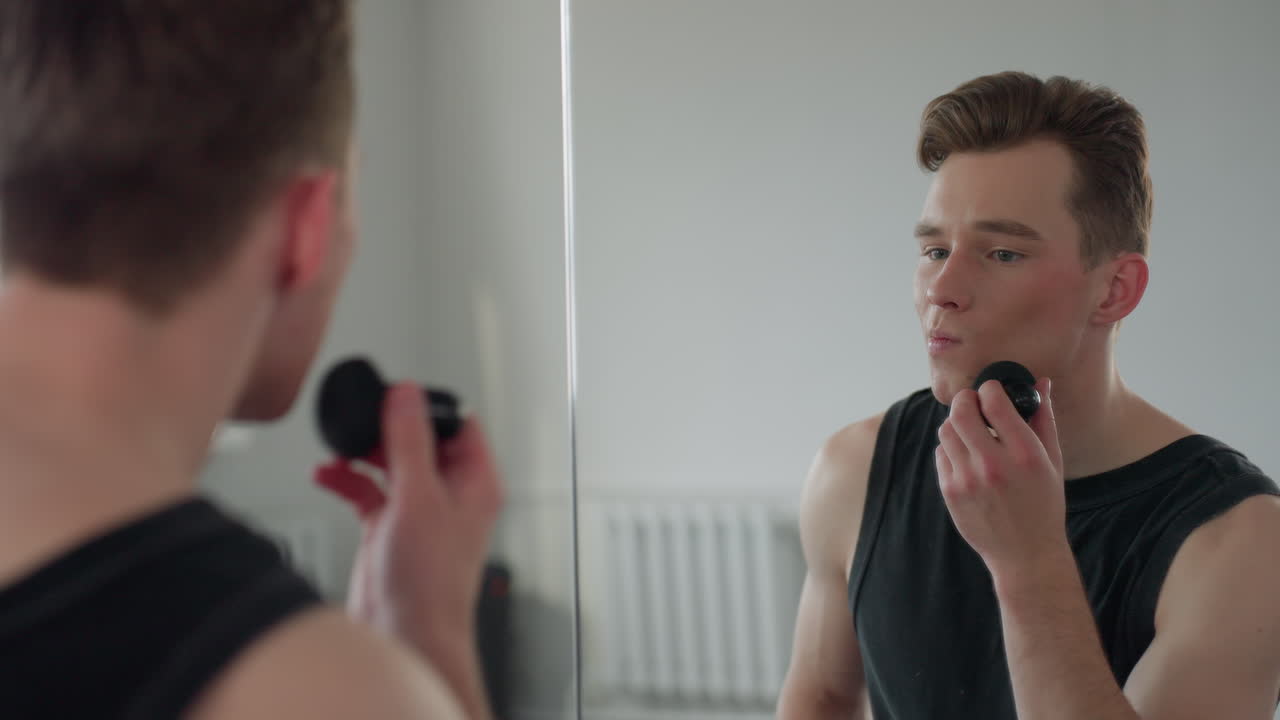 fair skinned boy in black vest applies makeup with sponge in front of large mirror, soft daylight fills minimalist studio with urban window view and reflection capturing grooming routine