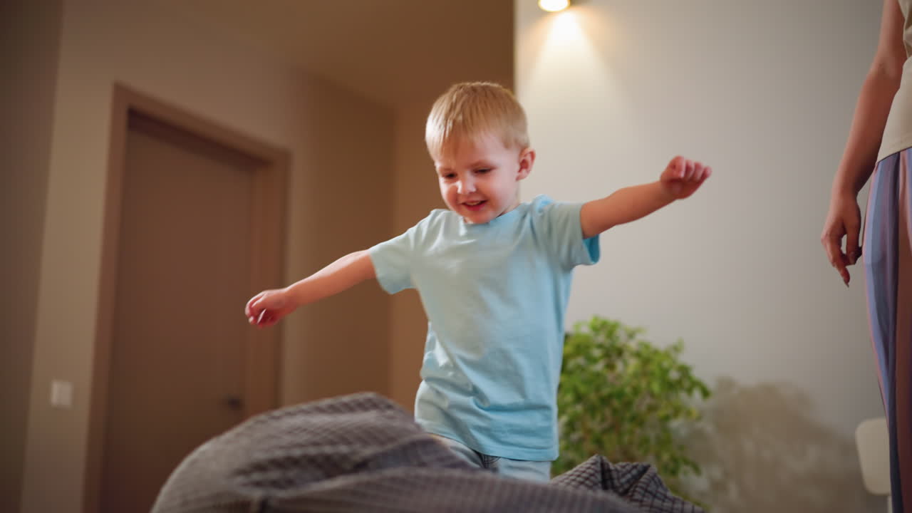 Child in light blue shirt spreads arms walking playfully indoors with smile, showing carefree joy, balance, and fun energy in cozy home environment with soft lighting, green plant, family atmosphere