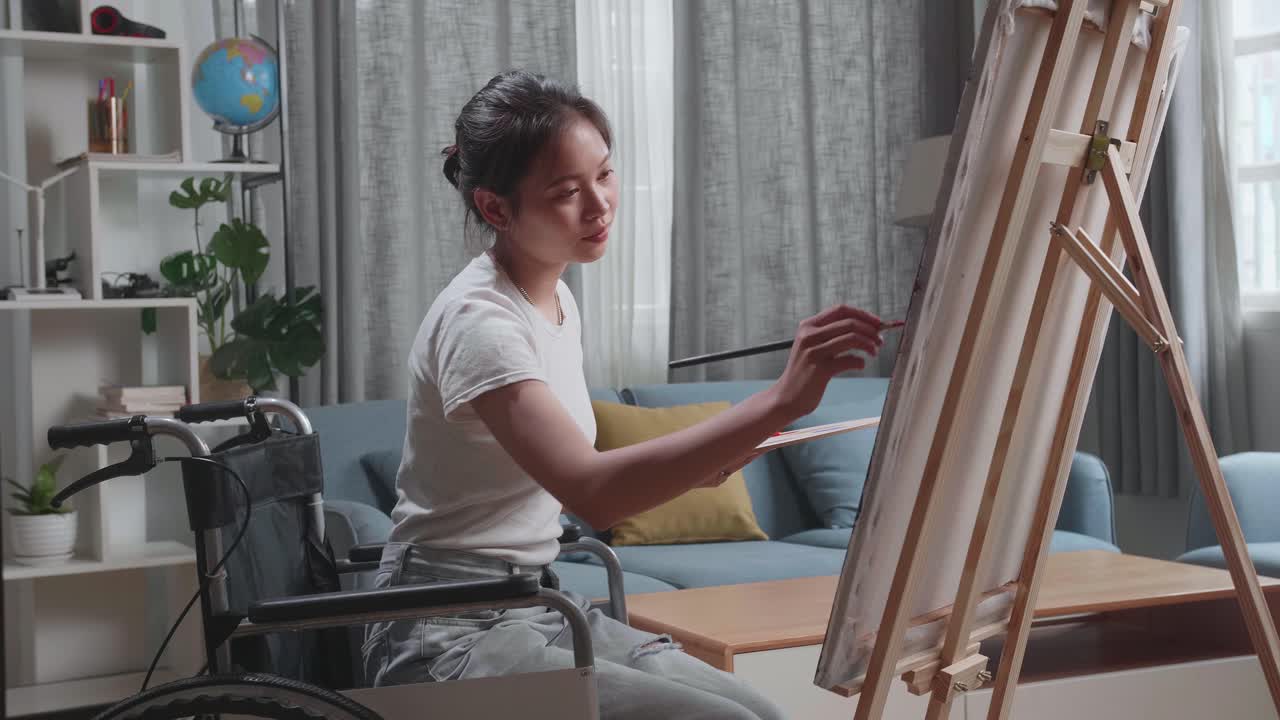 Medium Shot Side View Of An Asian Artist Girl In Wheelchair Holding Paintbrush Mixed Colour Thinking And Painting On The Canvas