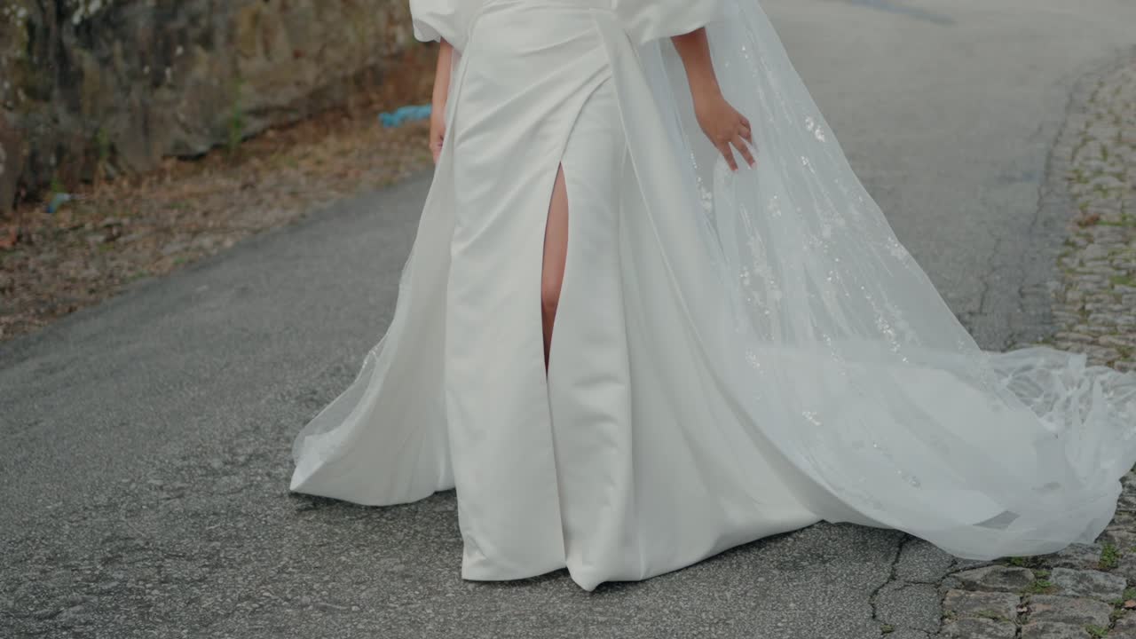 Bride walking in a white gown with a slit, holding the flowing fabric of her dress outdoors.