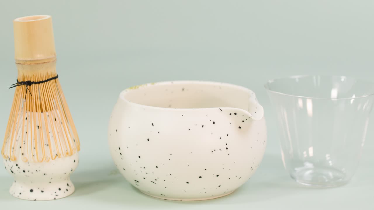 Clear water pours from glass bottle into ceramic bowl beside bamboo whisk, minimal clean setup