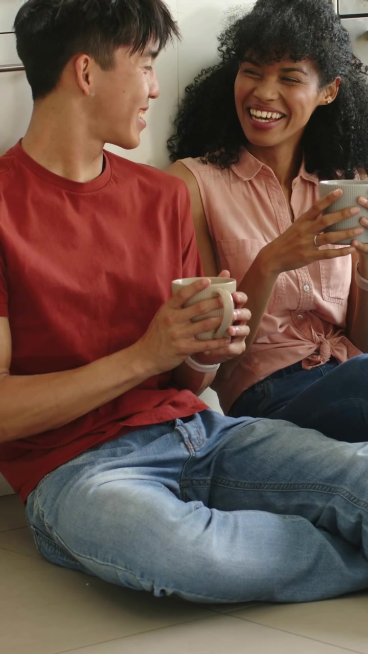 Video vertical: Pareja riendo bebiendo de tazas después de bromear en el suelo de baldosas de la cocina, mujer con top rosa.