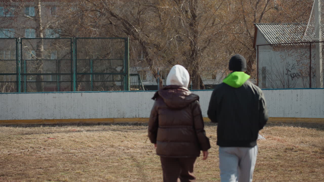 흰색 겉옷과 후드를 입고 동네 경기장 빈 골대를 향해 걸어가는 십 대 백인 아이들, 카메라를 등지고, 맑은 봄 날씨, 차분한 아파트 배경, 느린 걸음, 기대감과 집중된 시선