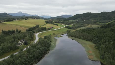 straight flying camera movement above the norwegian lake with beautiful reflections of the sky surrounded by mountains and forests as well as yellow and red houses as the camera passes by the road