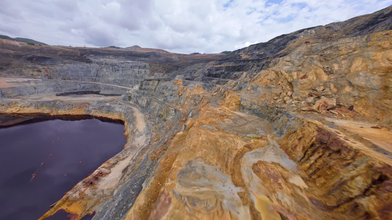 Stark landscape of open-pit gold and silver mine, colorful mineral-stained rock walls and a dark, contaminated tailings lake, Barrick Gold, Cotuí, Dominican Republic. Aerial drone FPV
