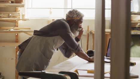 African american male carpenter talking on smartphone and using laptop in a carpentry shop