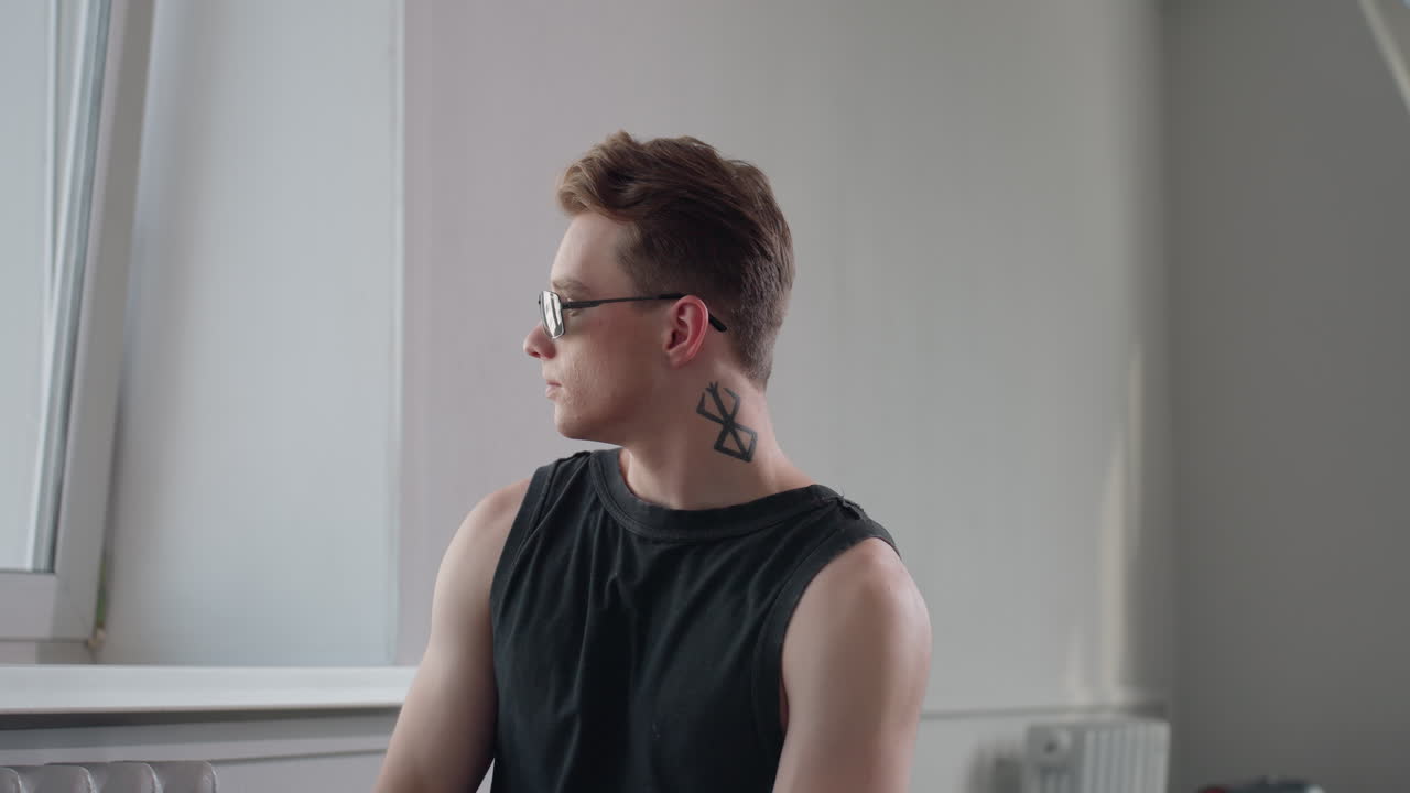 Stylish boy with neck tattoo indoors by tilted window puts on dark shades while gazing through glass in serene pose with soft light casting shadows on textured wall and radiator backdrop