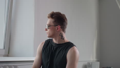 stylish boy with neck tattoo indoors by tilted window puts on dark shades while gazing through glass in serene pose with soft light casting shadows on textured wall and radiator backdrop