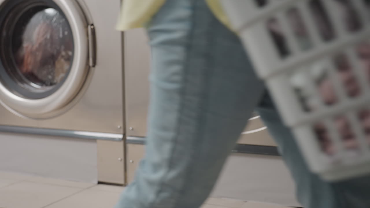 Interior washing machine drum as female hand removes fabric, drops into basket on floor, close view of towels, stainless rim, hygiene service workflow inside laundromat, clean process