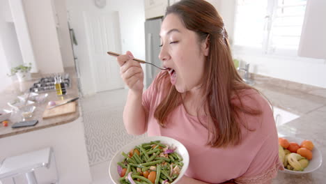 mujer biracial de tamaño plus feliz comiendo ensalada de verduras de pie en la cocina, cámara lenta