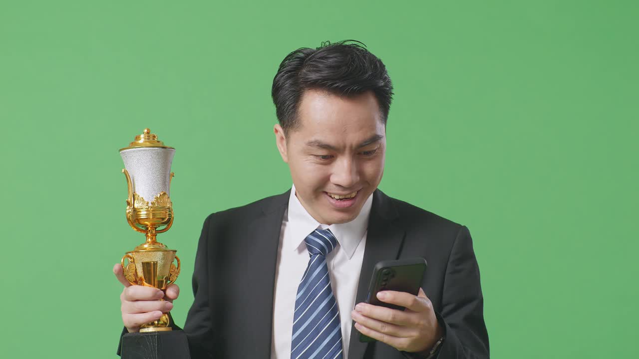 슈트와 타이를 입은 아시아 사업가, 금메달과 트로피를 들고 스마트폰을 쳐다보며 스튜디오의 녹색 스크린 배경에서 와우라고 말합니다.