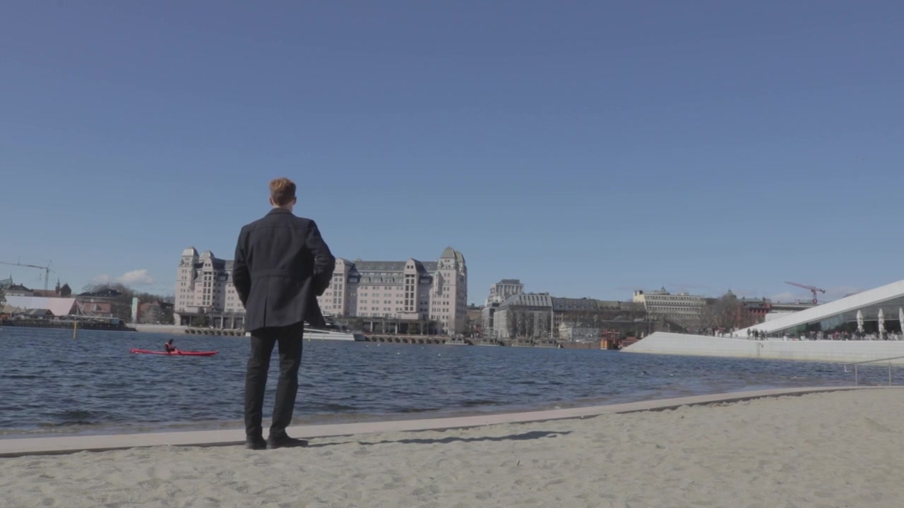 Uomo in piedi sulla spiaggia di Oslo vicino al Teatro dell'Opera, che si affaccia sul porto e sullo skyline della città.