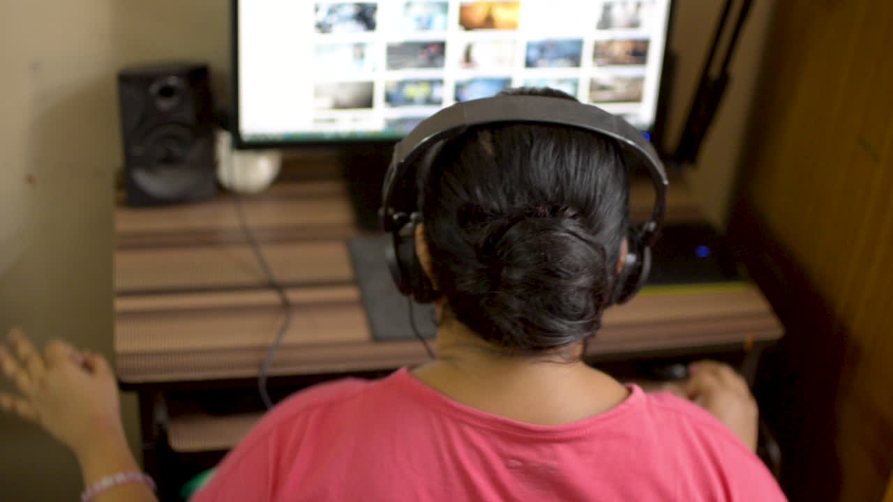 Indian asian caucasian woman girl in pink t-shirt listening enjoying music wearing headphone on desktop pc computer at home