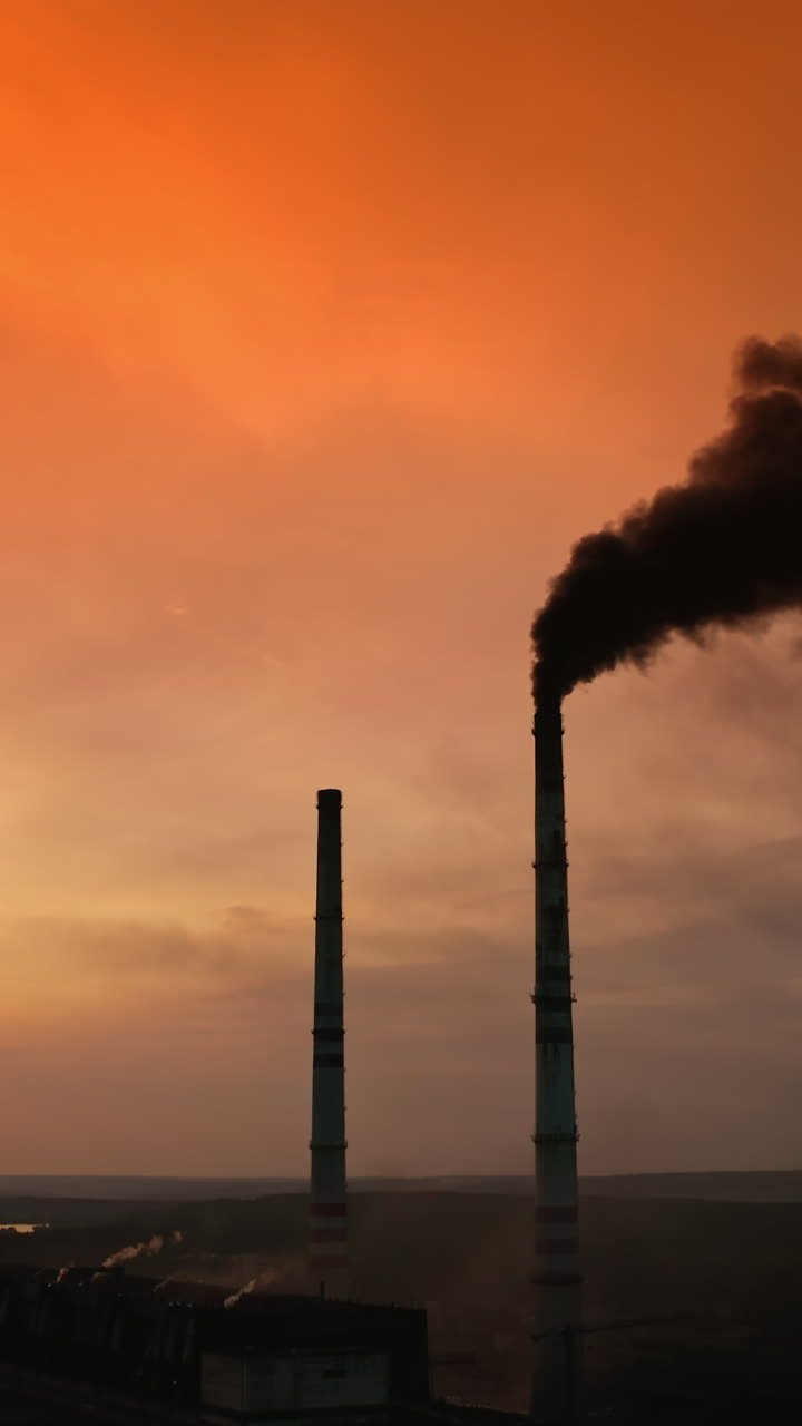 Black smoke from industrial pipe against orange sky. Silhouette of factory chimneys with dark fumes near the river at sunset. Chemical smoke pollute the environment. Ecological disaster. Vertical video