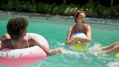laughing diverse female friends enjoying pool day, playing with beach ball and relaxing on float