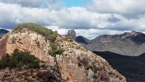 drone camera glides over rugged split rock, revealing dramatic cliffs, forested slopes, and distant peaks under partly cloudy daylight in a remote australian landscape