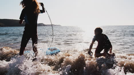 two silhouetted children wearing wetsuits playing in sea with bodyboards on  summer beach vacation