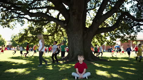 a young boy sits peacefully under a large tree while other children play in the schoolyard