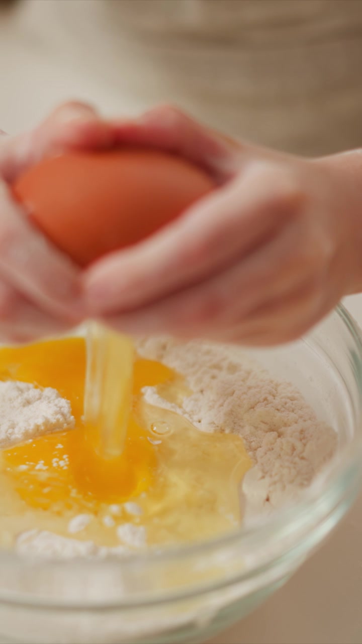Woman cracks egg into bowl of flour mixing ingredients for baking. close up view highlights texture of eggs and dry mixture in soft natural light against neutral background