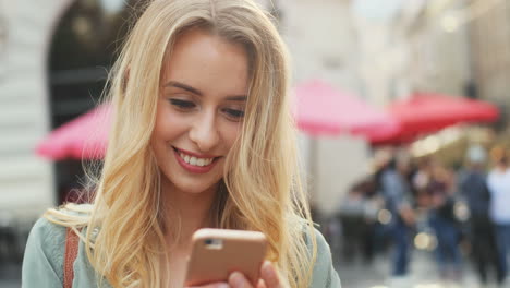 Vista-Cercana-De-La-Mujer-Rubia-Caucásica-Enviando-Mensajes-De-Texto-En-El-Teléfono-Inteligente-Y-Sonriendo-En-La-Calle