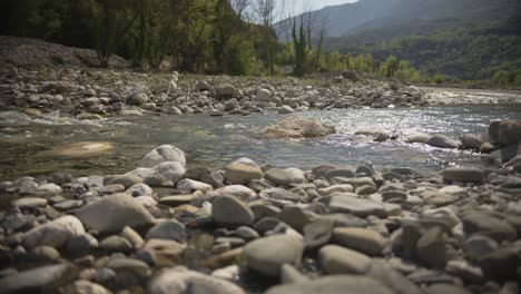 Smooth, sunlit stones in the foreground as the shallow waters of Arachthos River flow gently in the background. The surrounding landscape includes forested slopes and distant mountains of Tzoumerka.