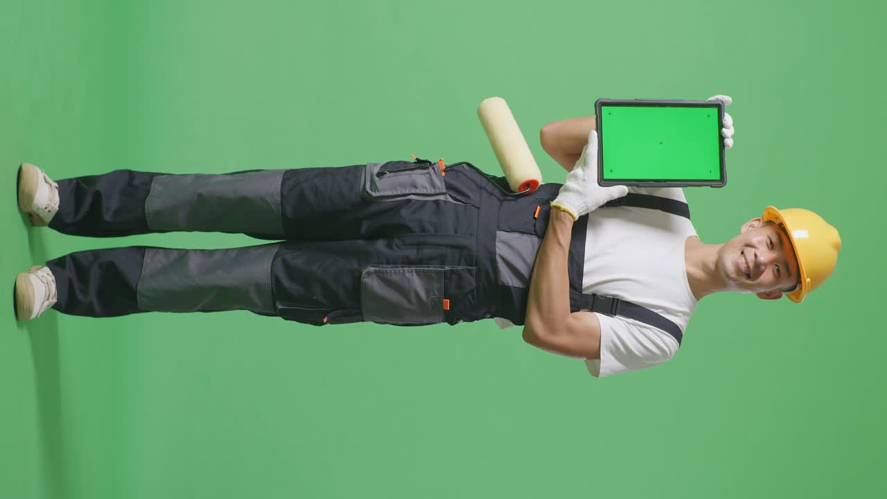 Full Body Of Asian Man Painter Wearing Safety Helmet Smiling And Showing Green Screen Tablet To The Camera While Standing In The Green Screen Background Studio