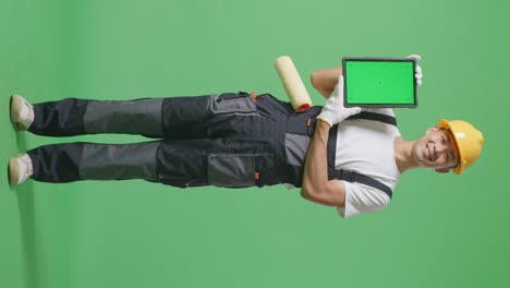 full body of asian man painter wearing safety helmet smiling and showing green screen tablet to the camera while standing in the green screen background studio