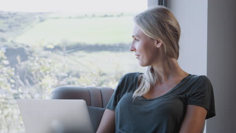 Mujer-Relajándose-En-Una-Silla-Junto-A-La-Ventana-En-Casa-Usando-Una-Computadora-Portátil