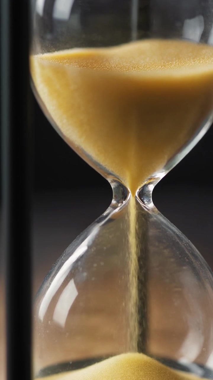 Glass hourglass with black frame on wooden table as golden sand trickles down