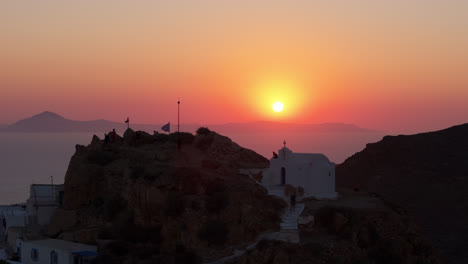aerial telephoto pan over anafi coast at golden hour with sun setting over serene landscape as greek flag waves and pink glow sets over water and white homes