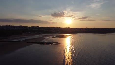 Sunset-rays-beam-fiery-golden-colours-across-relaxing-beach-ocean-coastline-aerial-view-rising-over-water-tide