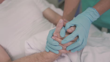 detail of a nurse's hands with gloves grasping and comforting the hands of a sick patient on hospital bed