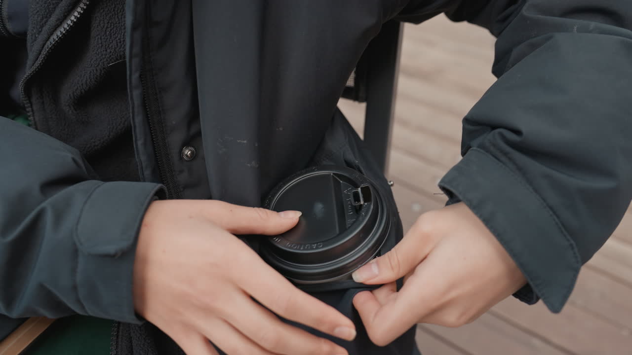 ガラスのテーブルに置かれたコーヒーカップ、テイクアウトカップを持ち上げて内ポケットにしまい込むためにジャケットに手を伸ばす、屋外ベンチの風景、涼しい朝、温かい飲み物を確保するカジュアルな通勤者、クローズアップの詳細