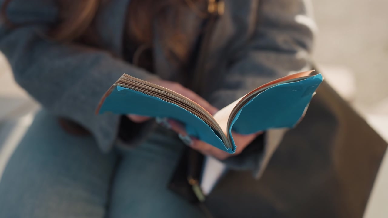 Bella donna pendolare caucasica con un diario turchese in grembo che sfoglia le pagine mentre aspetta all'aperto, con cappotto di jeans e grigio visibile, scena spontanea su panchina in città, carta tattile e copertina cucita, appunti di viaggio.