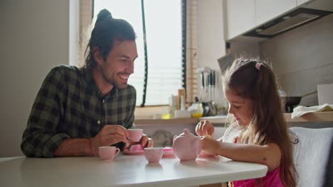 Un-Hombre-Moreno-Feliz-Con-Una-Camisa-A-Cuadros-Verde-Y-Su-Pequeña-Hija,-Una-Niña-Morena-Con-Un-Vestido-Rosa,-Juegan-A-Tomar-El-Té-Usando-Tazas-Rosas-De-Juguete-Y-Una-Tetera-En-La-Cocina