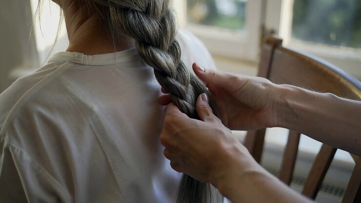 Le mani di una trecciatrice intrecciano capelli a casa, lisciando una maglietta bianca e preparando la punta con un elastico