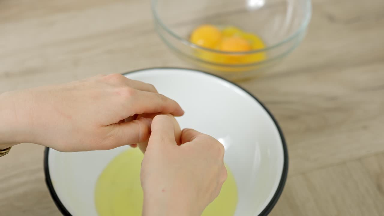Unrecognizable person cracks egg with a knife, separating yolk and white over a bowl for cooking, with visible yolks in glass bowl nearby, home kitchen environment with wood surface, making tiramisu