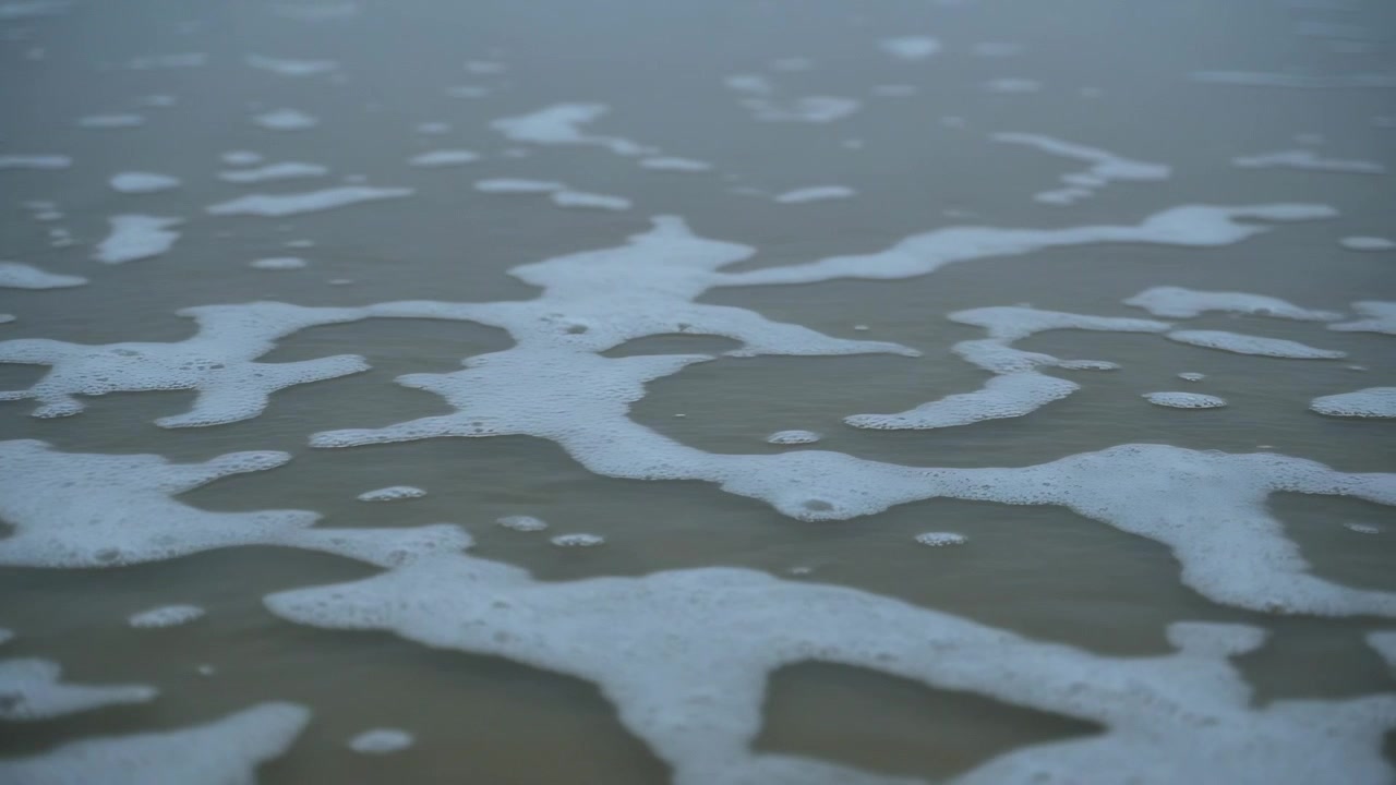 Shifting sea foam patches changing shape on wet sand at shoreline, thin seawater film moving