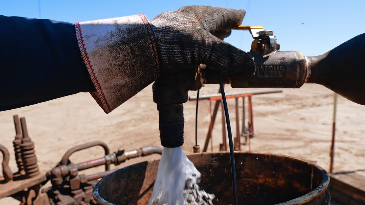 Male hand in a protective glove holding the tap on the pipe. White liquid is poured from the faucet under the pressure. Industrial drilling setting.